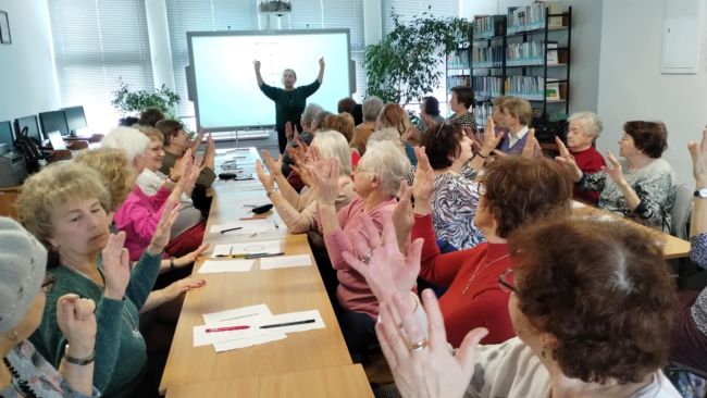 Seniorzy siedzą przy stołach i wykonują ćwiczenia oddechowe i palcowe poprawiające koncentrację.