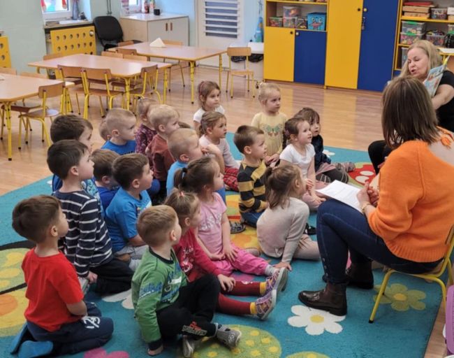 Nauczyciele czytają książkę dzieciom, które siedzą na dywanie i słuchają.