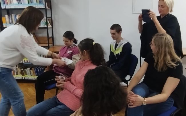 Uczestnicy zajęć siedzą na krzesłach, nauczyciel rozdaje niespodzianki.