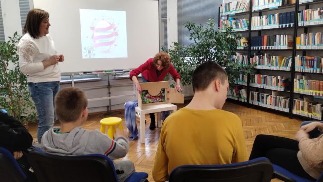 Nauczyciele czytają opowiadania, dzieci siedzą na krzesłach i słuchają.
