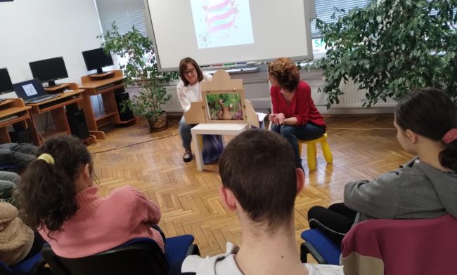 Nauczyciele czytają opowiadania, dzieci siedzą na krzesłach i słuchają.