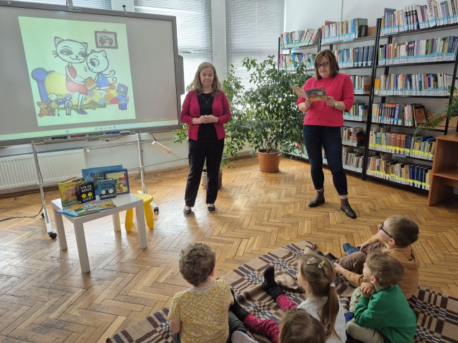 Nauczyciele stoją i prezentują bajkę na tablicy, dzieci siedzą na podłodze i słuchają.