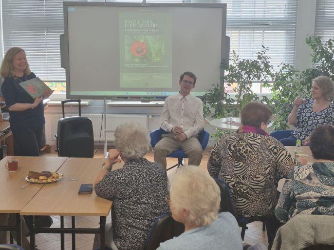 Spotkanie w czytelni Biblioteki Pedagogicznej. Uczestnicy siedzą przy stolikach, autorzy wystawy i prowadząca w centrum siedzą na fotelach, prowadzą rozmowę.