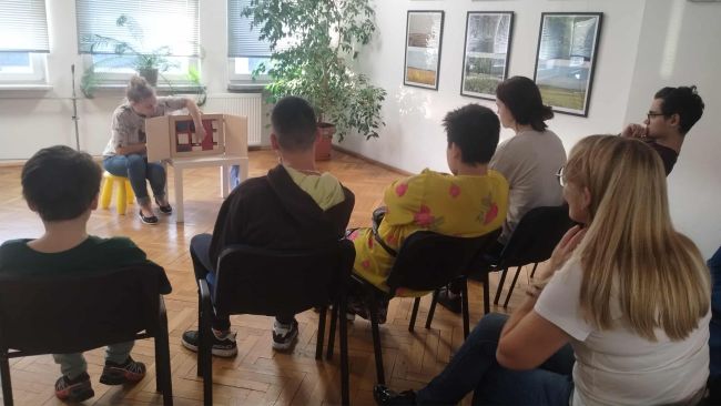 Uczniowie siedzą na krzesłach. Słuchają opowiadania.