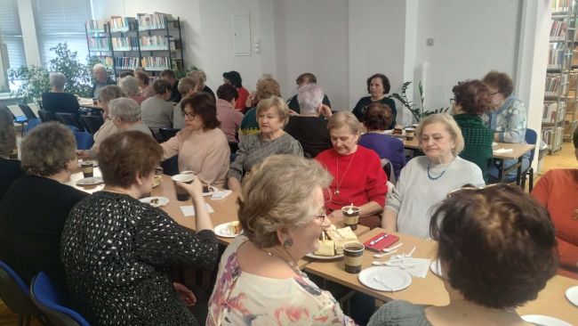 Uczestnicy wydarzenia siedzą przy stolikach. Częstują się kawą i ciastkami. W tle, zaproszenie goście siedzą na fotelach i prowadzą spotkanie.