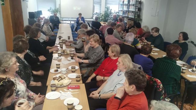 Uczestnicy wydarzenia siedzą przy stolikach. Częstują się kawą i ciastkami. W tle, zaproszenie goście siedzą na fotelach i prowadzą spotkanie.