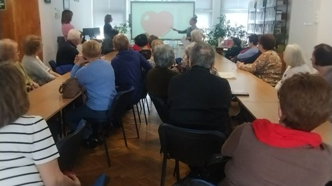 Słuchacze Uniwersytetu Trzeciego Wieku z Tomaszowa Lubelskiego siedzą przy stolikach, uczestniczą w zajęciach. Prowadzące stoją przy tablicy interaktywnej, tłumaczą wykonanie zadania.