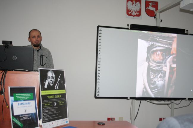 Na zdjęciu widoczny prelegent Andrzej Maziarz przy mównicy w czytelni biblioteki pedagogicznej, a na tablicy interaktywnej slajd z jego prezentacji prezentujący astronautę