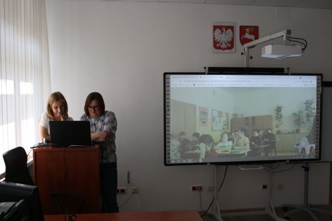 Dwóch nauczycieli bibliotekarzy Biblioteki Pedagogicznej w Zamościu w tle tablica multimedialna z wyświetloną klasą - uczestnikami warsztatów online