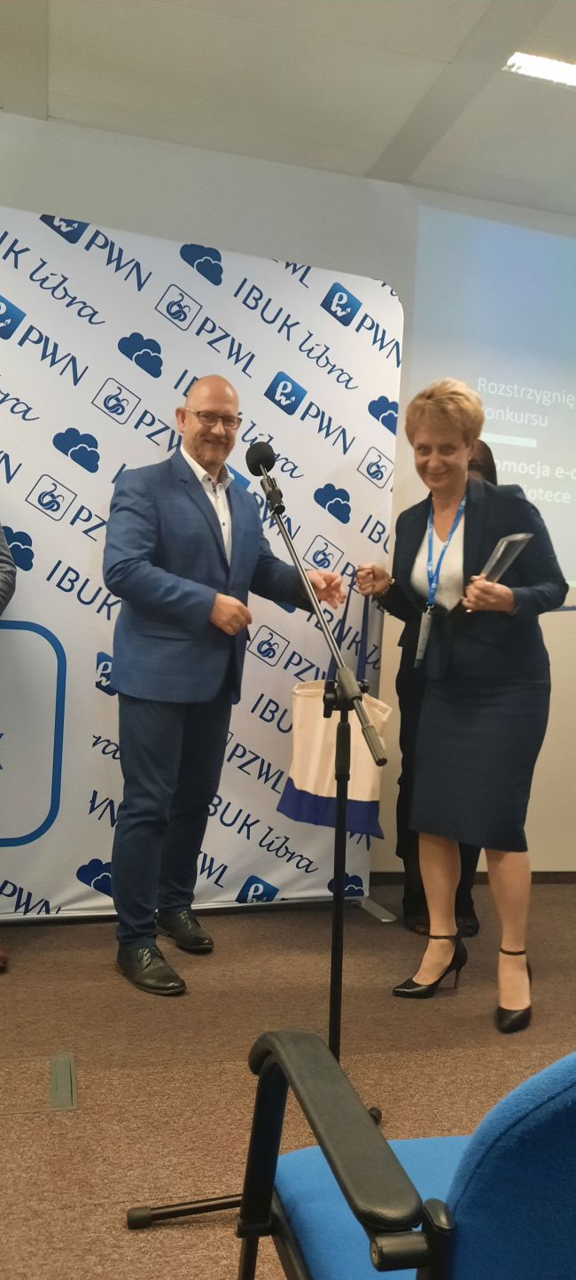 Na zdjęciu po lewej stronie znajduje się Pan Piotr Kołacz, wręczający statuetkę za zajęcie I miejsca Pani Dorocie Mach-dyrektor Biblioteki Pedagogicznej w Zamościu. Obie postaci pozują na tle rozwijanego banneru-typu rooll-up z logo PWN, Ibuk Libra i PZWL.