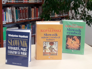 Na zdjęciu zaprezentowane są trzy książki Władysława Kopalińskiego "Słownik wydarzeń, pojęć i legend XX wieku", "Słownik mitów i tradycji kultury" oraz "Słownik symboli". Książki są ustawione na stoliku w czytelni Biblioteki Pedagogicznej w Zamościu, w tle widać regał z książkami i wysoki, rozłożysty kwiatek doniczkowy.