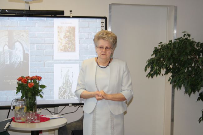 Na zdjęciu widać Marię Duławską. Po lewej na stoliku bukiet kwiatów, a w tle na tablicy interaktywnej wyświetlają się okładki książek Marii Duławskiej.