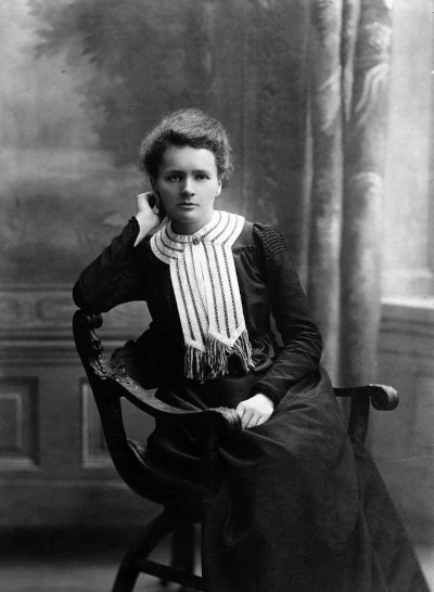 Czarno-białe zdjęcie przedstawia Marię Curie-Skłodowską w 1903 roku w wieku 36 lat. Siedzi na krześle, jest ubrana w długą ciemną suknię z ozdobnym jasnym kołnierzykiem na guzik przypominającym szalik.