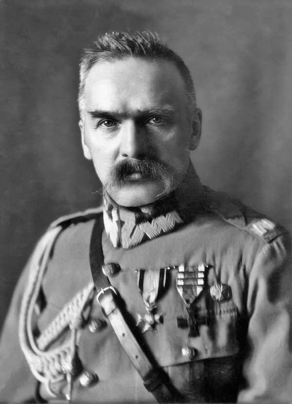 Czarno-białe zdjęcie przedstawia Józefa Piłsudskiego ubranego w mundur wojskowy, z przypiętymi odznaczeniami na lewej piersi. Jest krótko ostrzyżony, ma sumiaste wąsy.