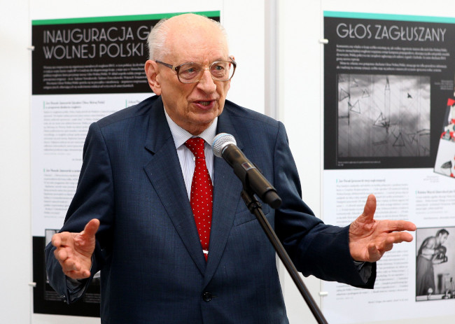 Zdjęcie przedstawia Władysława Bartoszewskiego w 2012 roku w wieku 90 lat. Stoi przed mikrofonem, przemawia. Jest ubrany w granatowy garnitur i czerwony krawat w drobne kropki. Na nosie ma spore okulary o brązowych oprawkach.