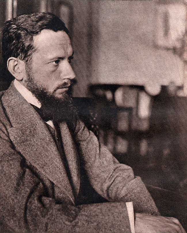 Zdjęcie przedstawia Leopolda Staffa około roku 1910. Poeta siedzi, jego sylwetka jest ujęta z profilu. Prezentuje się jako szczupły mężczyzna, jest ubrany w marynarkę, ma sporą brodę, włosy niezbyt długie.