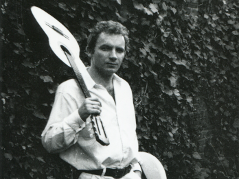 Czarno-białe zdjęcie Edwarda Stachury ze zbiorów Muzeum Literatury im. Adama Mickiewicza. Na zdjęciu Edward Stachura stoi na tle wysokiego żywopłotu, prawą ręką trzyma gitarę na swoim prawym ramieniu, w lewej ręce trzyma kapelusz. Jest ubrany w jasną koszulę i jasne spodnie z czarnym paskiem.