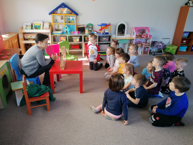 Na zdjęciu widać Joannę Magdziak, nauczyciela bibliotekarza Biblioteki Pedagogicznej, podczas zajęć w przedszkolu. Na zdjęciu widać również dzieci, które siedzą na wykładzinie i czekają na otwarcie teatrzyku kamishibai. W tle widać książeczki i zabawki dziecięce.