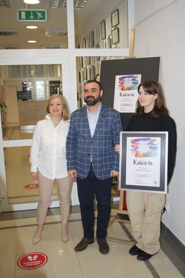Zdjęcie z otwarcia wystawy rysunku i malarstwa Wiktorii Zawrotniak. Na zdjęciu od lewej: wicedyrektor Biblioteki Pedagogicznej w Zamościu Anetta Sienkiewicz, dyrektor SP nr 6 w Zamościu Sebastian Plewa oraz autorka wystawy Wiktoria Zawrotniak trzymająca w rękach pamiątkowy plakat wystawy.
