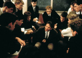 Zdjęcie prezentuje kadr z filmu Stowarzyszenie Umarłych Poetów. Na zdjęciu widać Johna Keatinga, granego przez Robina Williamsa, który przykucnął przy stolika swoich uczniów i opowiada im o tym, dlaczego warto poznawać literaturę. Uczniowie słuchają z zaciekawieniem. Wszyscy ubrani są w koszule z krawatami, większość ma też na sobie marynarki.