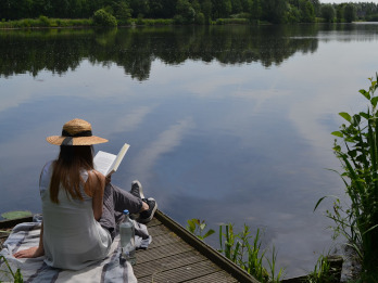 Na zdjęciu widać od tyłu kobietę, która siedzi na kocu na mostku nad jeziorem i czyta książkę. Ma na głowie słomiany kapelusz. W tle widać jezioro, a dookoła niego drzewa.