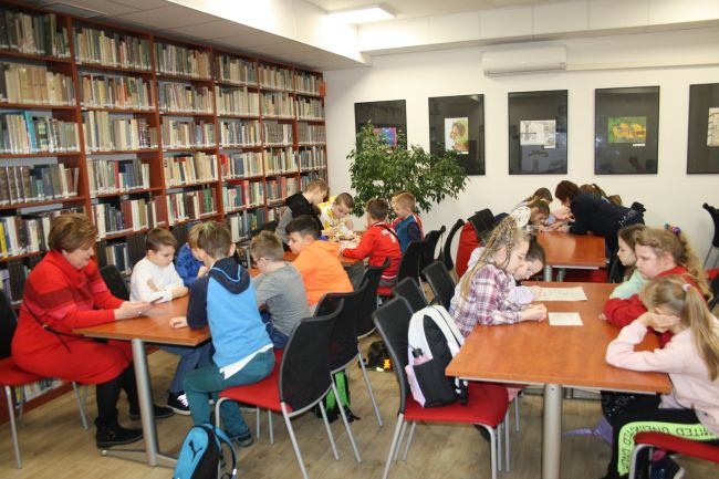 Na zdjęciu ukazani są uczestnicy warsztatów poświęconych Marii Konopnickiej, którzy przy stolikach rozwiązują zadania w ramach pokoju zagadek.