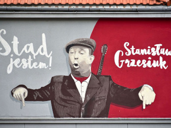 Zdjęcie prezentuje mural upamiętniający Stanisława Grzesiuka przy ul. Lubkowskiej 7/9 w Warszawie. Na muralu widać Grzesiuka ubranego w berecik, za jego plecami wystaje bandżola. Grzesiuk palce wskazujące obu dłoni ma skierowane w dół, a na muralu widnieje napis "Stąd jestem!"