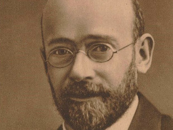 Czarno-białe zdjęcie przedstawia Janusza Korczaka. Na zdjęciu widać popiersie pedagoga. Jest ubrany w marynarkę, garnitur, krawat. Na nosi ma okulary z okrągłymi szkłami. Posiada brodę i łysą głowę. Spojrzenie ma życzliwe.