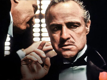 Zdjęcie przedstawia kadr z filmu "Ojciec chrzestny" Francisa Forda Coppoli. Widać na nim Vita Corleone, odgrywanego przez Marlona Brando. Jeden ze współpracowników szepcze mu coś na ucho. Vito Corleone ma wąsy, jest ubrany w garnitur i muszkę, palec wskazujący prawej ręki ma uniesiony lekko do góry.