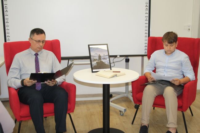 Nauczyciel oraz uczeń Katolickiej Szkoły Podstawowej czytają fragmenty Kordiana Juliusza Słowackiego. Na stoliku, przy którym osoby czytają, znajduje się w przezroczystej ramce wydrukowany obraz 