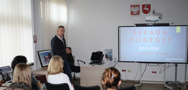 Jacek Podkościelny opowiada o swoich podróżach, na zdjęciu widać również część publiczności oraz tablicę multimedialną, na której wyświetla się napis 