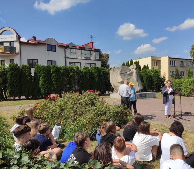 Na pierwszym planie młodzież siedzaca, w tle kobieta mówiąca do mikrofonu