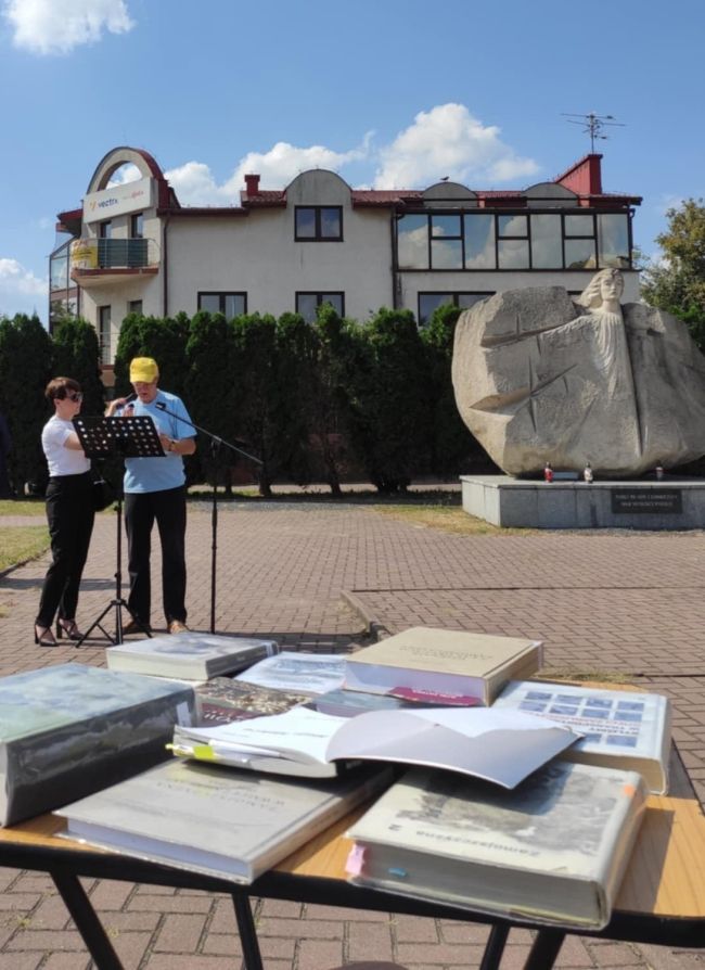 Na pierwszym planie ksiazki w tle mężczyzna czytający do mikrofonu
