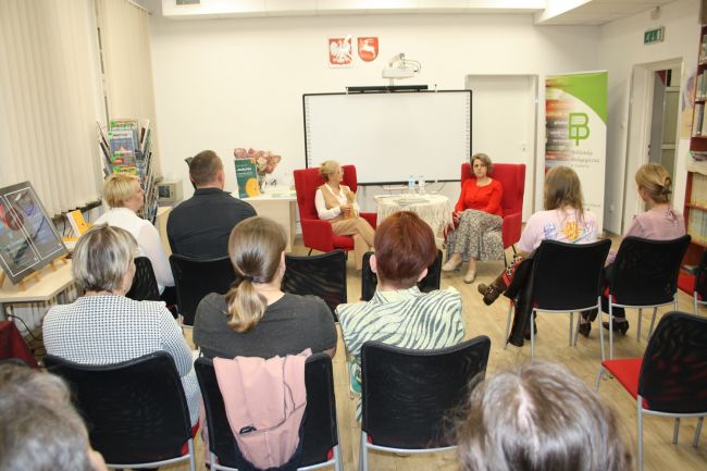 Wicedyrektor biblioteki oraz Monika Tadra podczas spotkania autorskiego. Zdjęcie zrobione z dalszej odległości, więc widać także siedzącą publiczność zwróconą w stronę prowadzącej oraz pisarki.