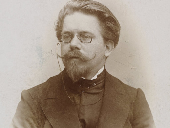 Fotografia przedstawia Władysława Reymonta. Jest ubrany w marynarkę i pod spodem długą kamizelkę pod szyję. Ma lekką bródkę i wąsy zawinięte po bokach. Włosy zaczesane do tyłu. Na nosie małe zaokrąglone okulary. Patrzy lekko w bok, w swoją prawą stronę.