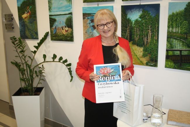 Regina Grzybowska na tle swoich obrazów prezentowanych na wystawie w Bibliotece Pedagogicznej.
