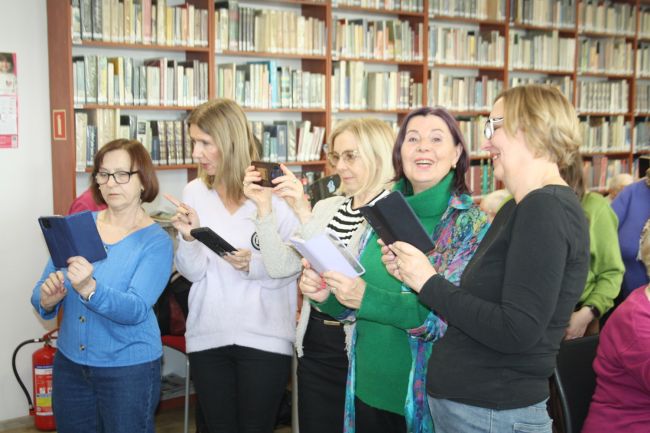 Uczestnicy zajęć siedzą przy stołach w czytelni Biblioteki Pedagogicznej, wiele osób się uśmiecha