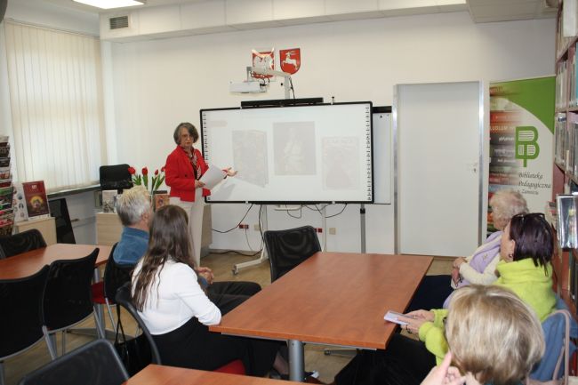 Dyrektor biblioteki dr Dorota Mach wygłasza prelekcję na temat kobiet w literaturze i malarstwie, której przysłuchują się seniorzy zebrani na kolejnym spotkaniu z cyklu