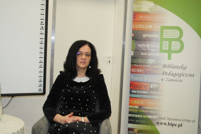 Prof. Sabina Bober podczas spotkania autorskiego, siedzi na tle roll-upu Biblioteki Pedagogicznej w Zamościu.
